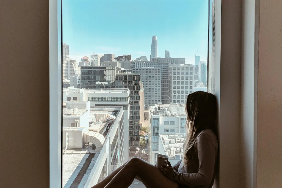 Woman sitting on a windowsill holding a mug, looking out over the city in a moment of reflection